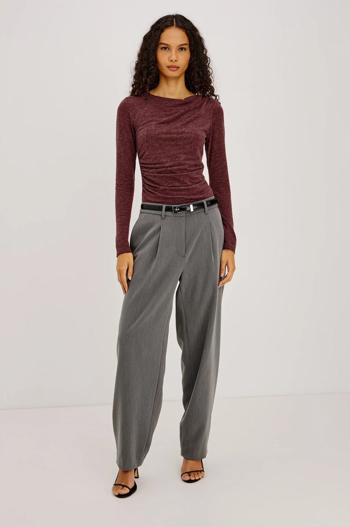 model wearing a long sleeve merlot top with a cowl neck and grey pants on a light grey background 