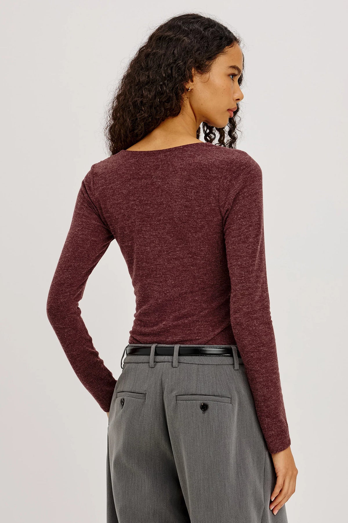 model wearing a long sleeve merlot top with a cowl neck and grey pants on a light grey background, back 