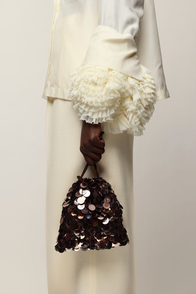 A close-up of a person wearing a cream-colored outfit with dramatic ruffled cuffs and holding Gala Bag in Dark Clove