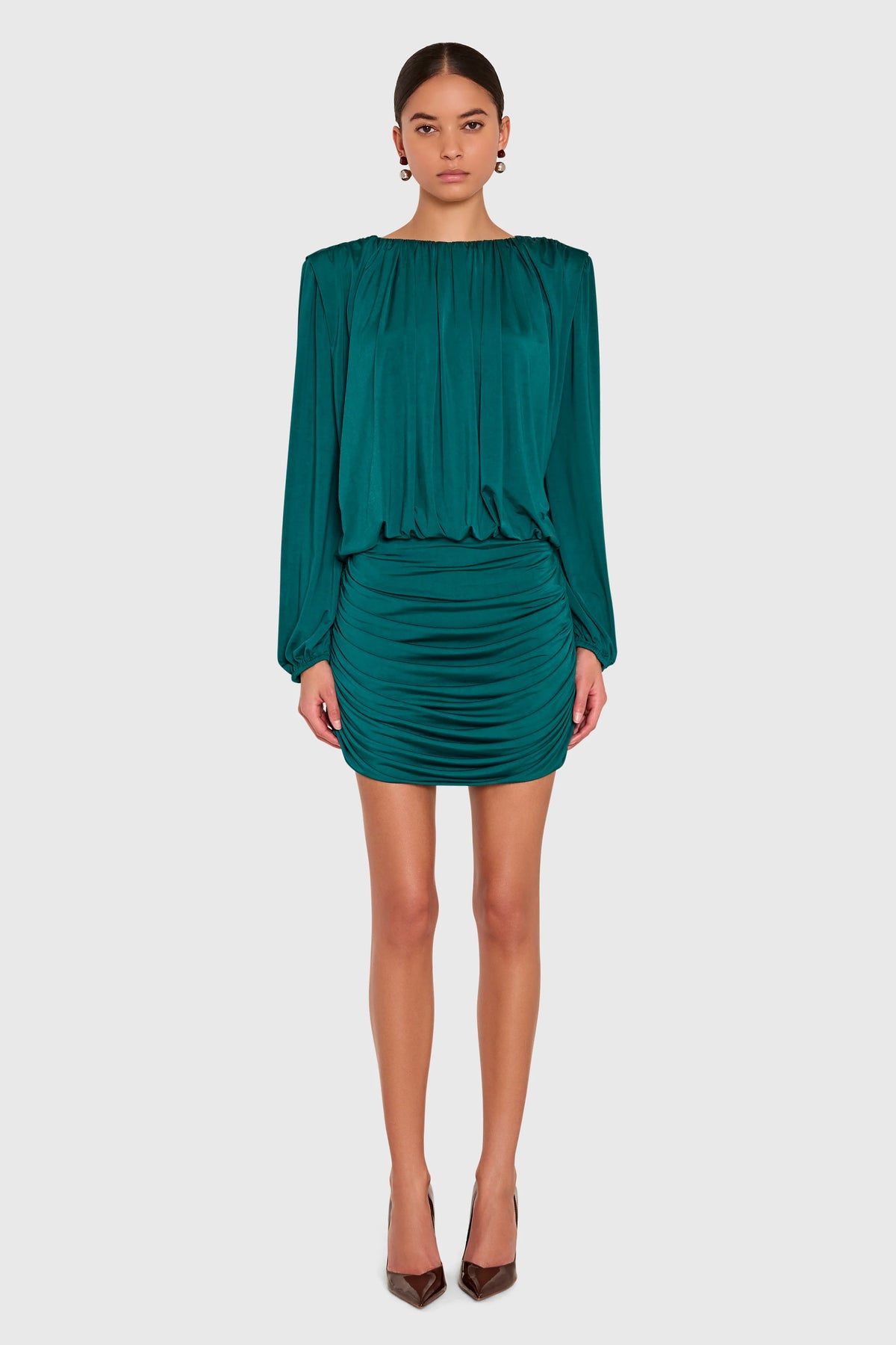 A model wearing a dark teal green long-sleeve mini dress with a gathered, pleated design, paired with brown pointed-toe heels, against a solid light gray background.