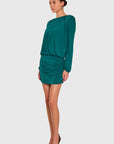 A woman modeling a teal green long-sleeve mini dress with pleated details on the bodice and a ruched mini skirt, paired with brown pointed-toe heels against a light gray studio background.