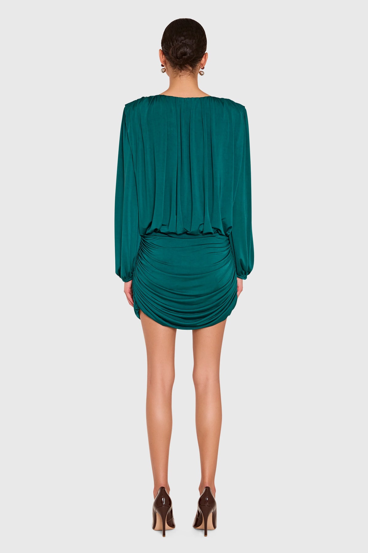 A woman shown from the back wearing a dark teal green draped mini dress with long sleeves and ruched detailing, paired with brown high heels, against a light gray studio background.