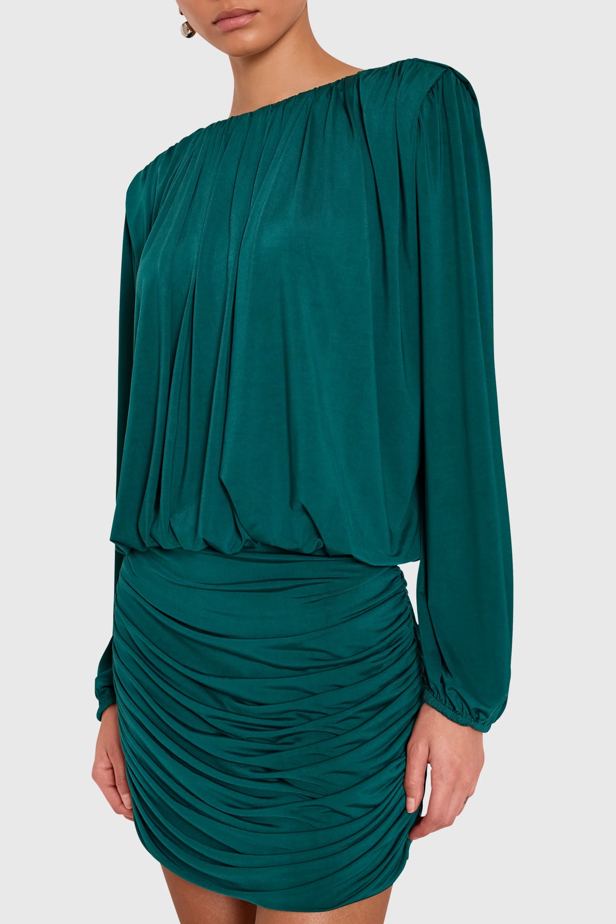 A woman wearing a dark teal green off-the-shoulder long-sleeve mini dress with ruched draping details, shown from the neck down against a light gray studio background.