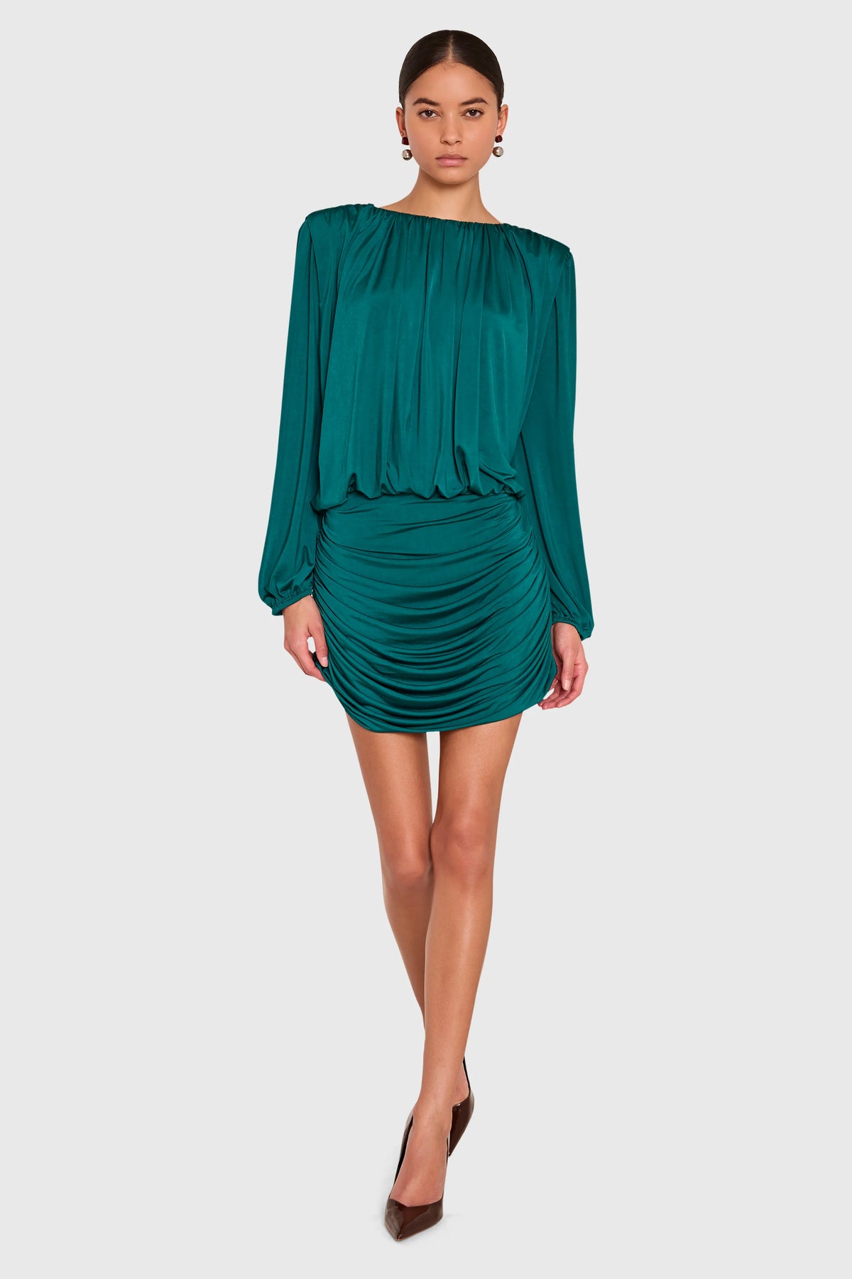 A model wearing a teal green off-the-shoulder long-sleeve mini dress with ruched detailing, paired with brown pointed-toe heels, against a solid light gray background.