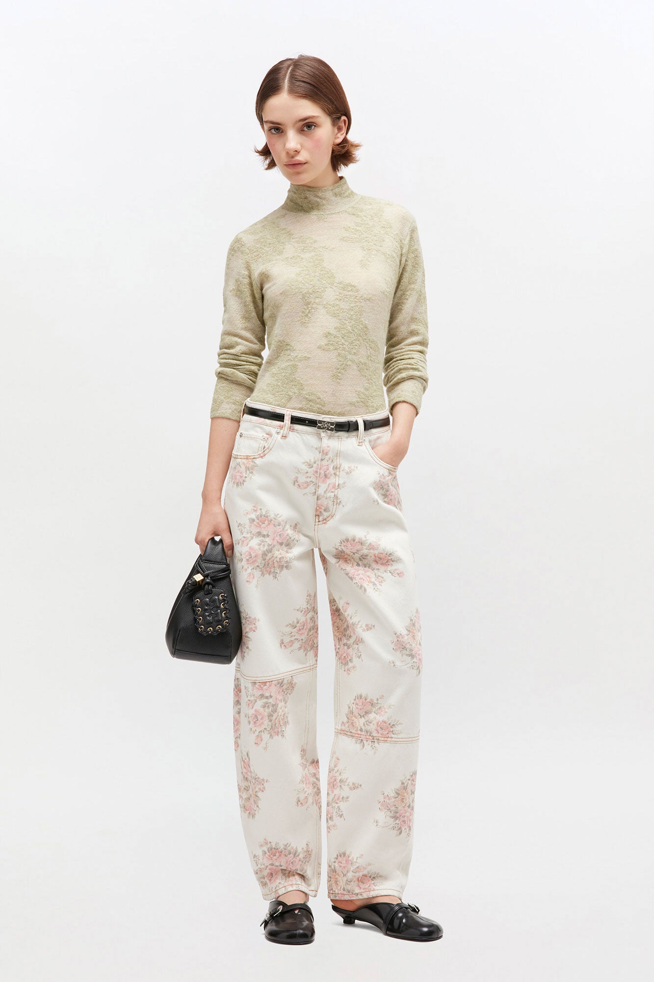 Woman wearing a light green sweater and floral pants with a black handbag on a white background