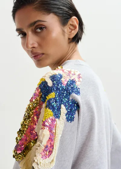 Woman wearing a gray sweatshirt with colorful sequin embellishments on the shoulders against a white background
