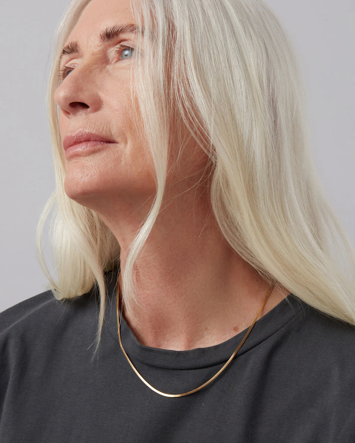 Person with blonde hair wearing a dark shirt and gold necklace against a gray background