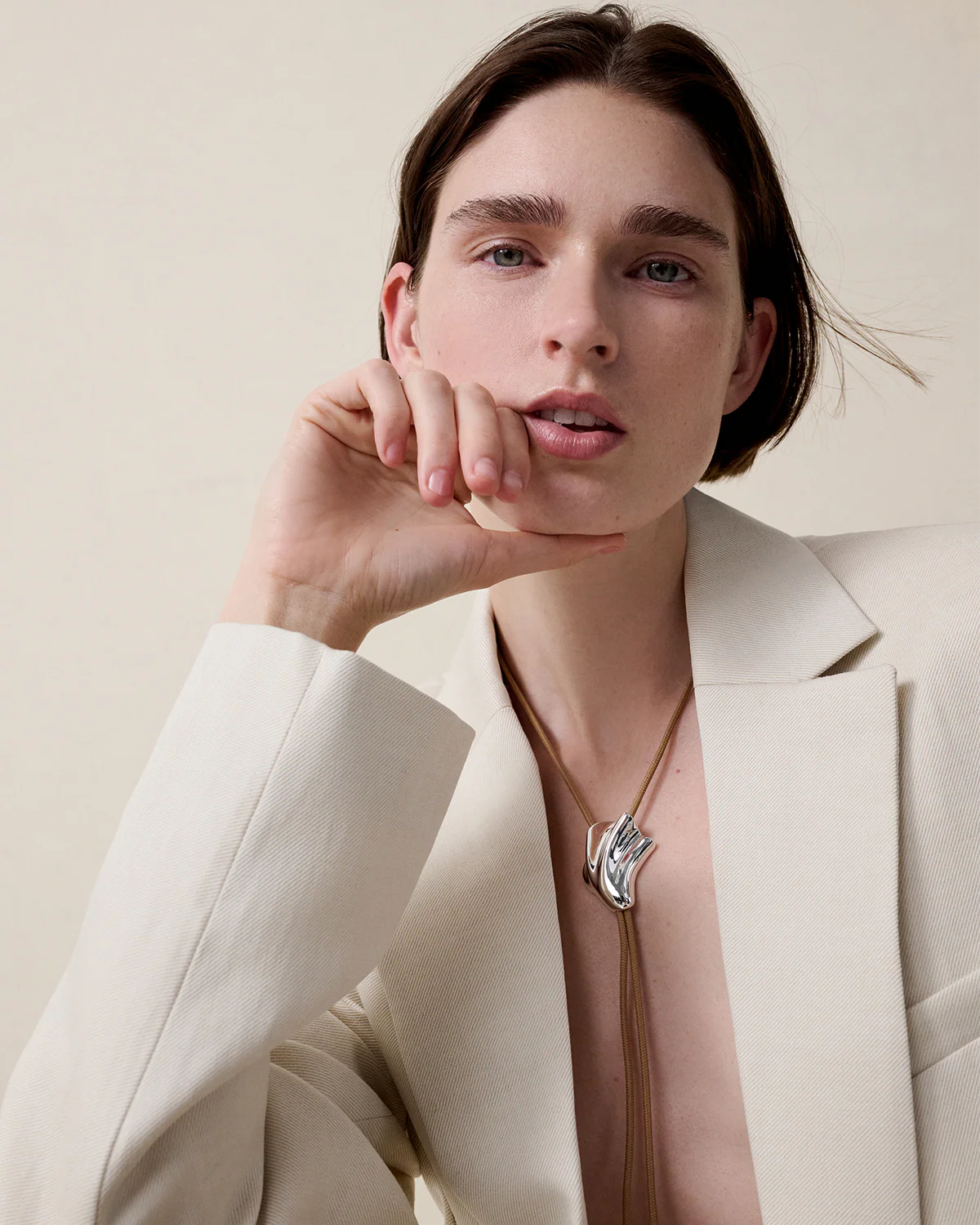 Woman wearing a beige blazer and a necklace with a bird design on a neutral background