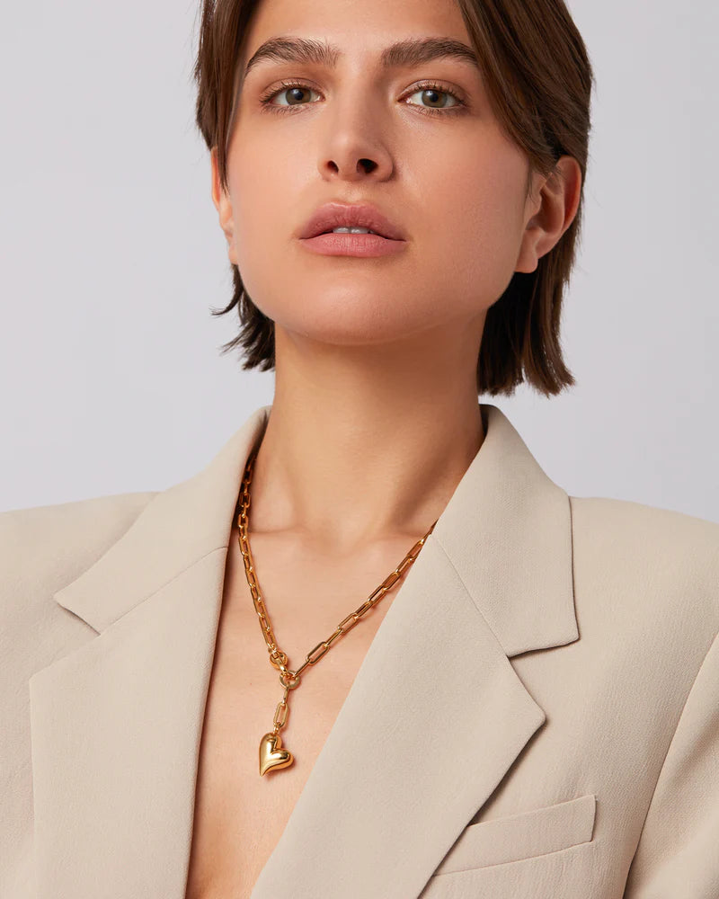Woman wearing a beige blazer and gold necklace with a heart pendant on a plain background