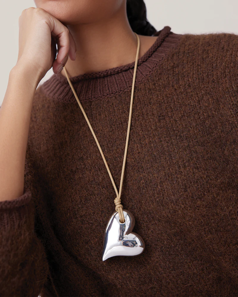Person wearing a brown sweater with a silver heart-shaped pendant on a long chain.