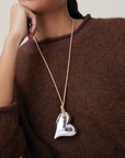 Person wearing a brown sweater with a silver heart-shaped pendant on a long chain.