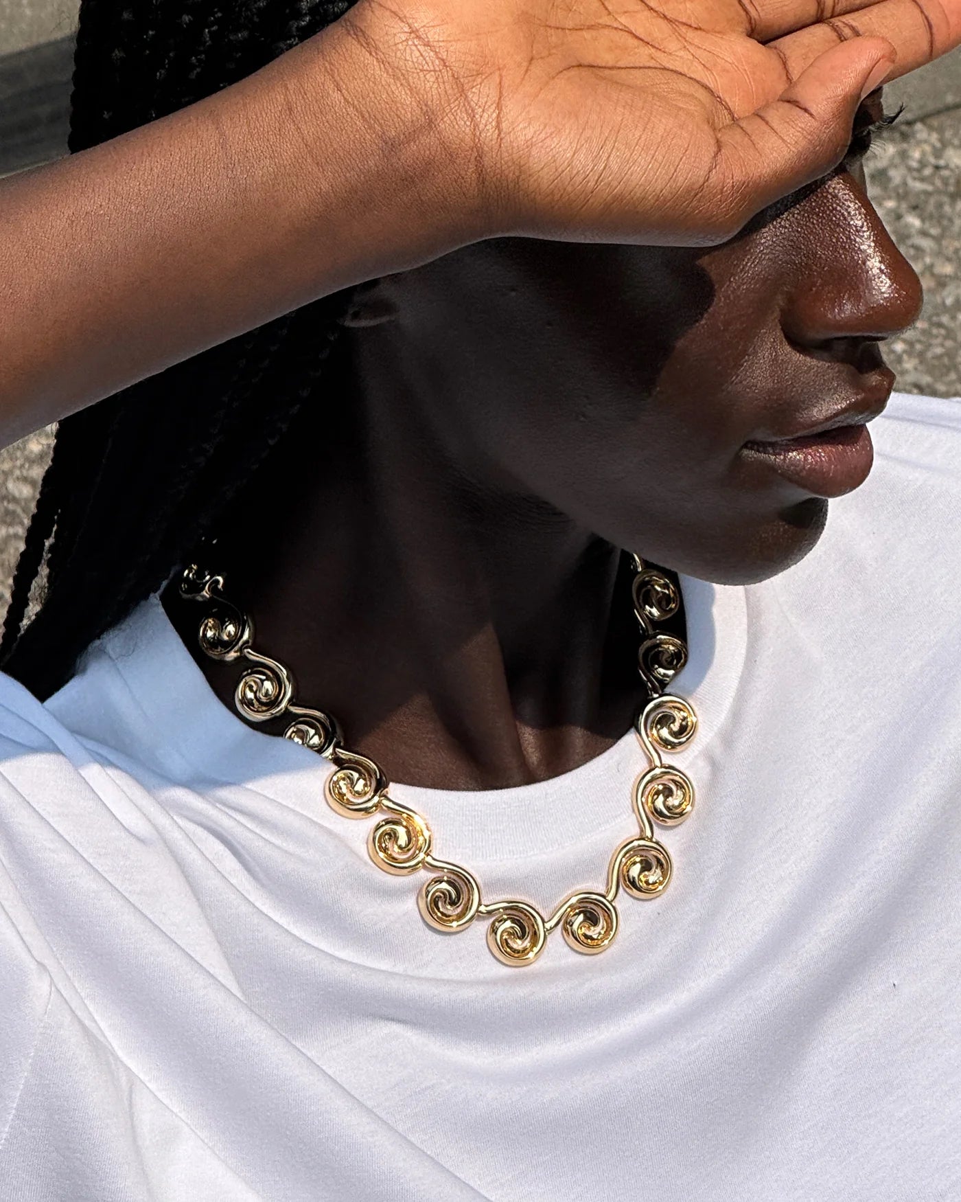 A close-up lifestyle shot of a person wearing a chunky gold chain necklace with a distinctive spiral or scroll link design, paired with a white t-shirt.