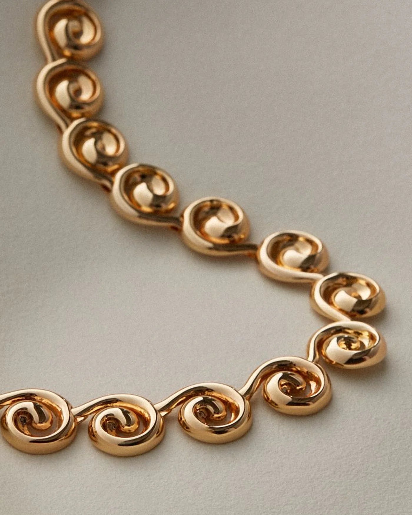 A close-up of a gold chain necklace featuring repeating polished spiral or scroll-shaped links, photographed on a plain light gray background.