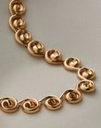 A close-up of a gold chain necklace featuring repeating polished spiral or scroll-shaped links, photographed on a plain light gray background.