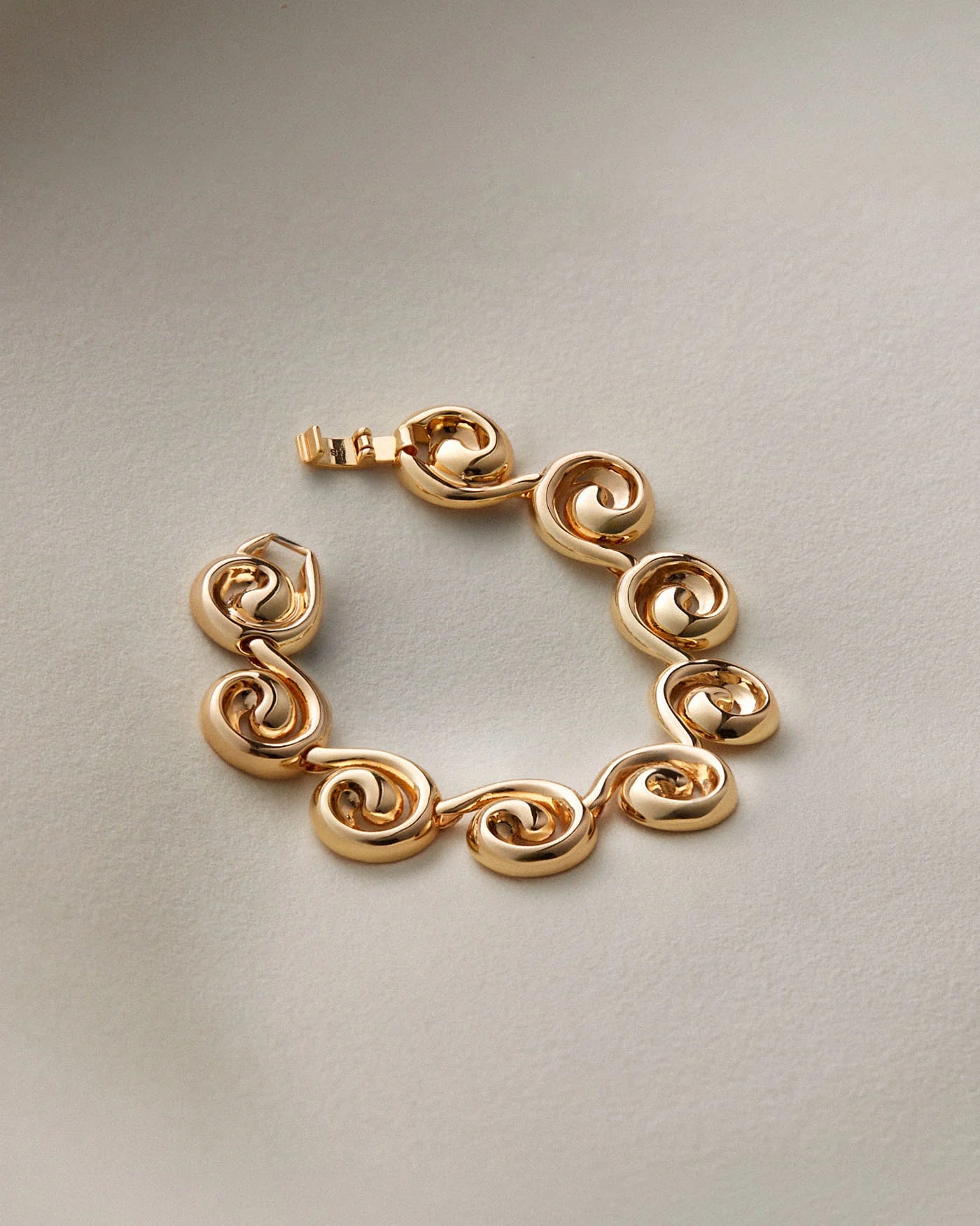 A gold-plated link bracelet with a unique spiral or scroll design on each link, displayed on a neutral light gray background.