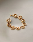 A gold-plated link bracelet with a unique spiral or scroll design on each link, displayed on a neutral light gray background.