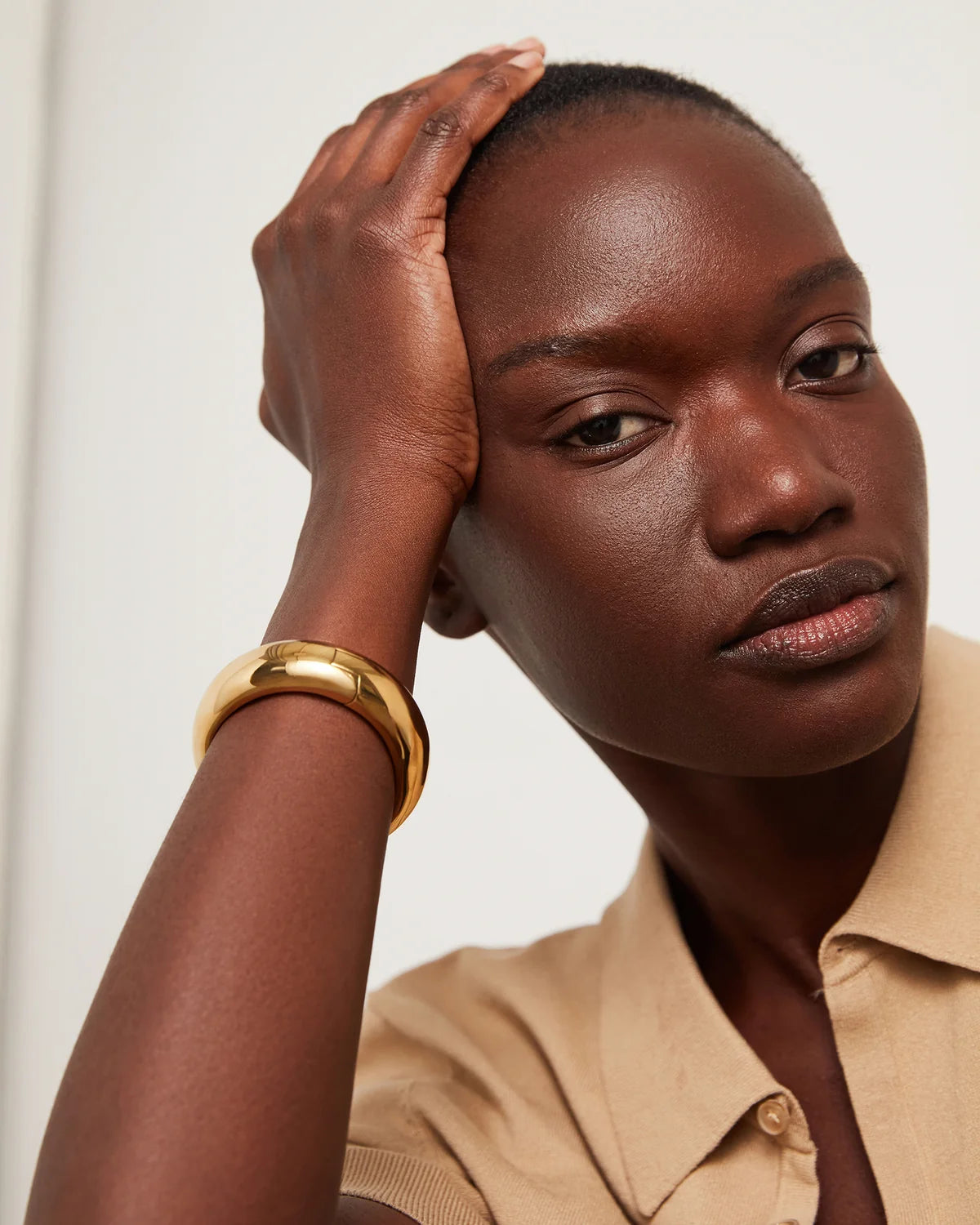 A close-up of a model wearing a beige with a gold bangle bracelet, against a plain light background.