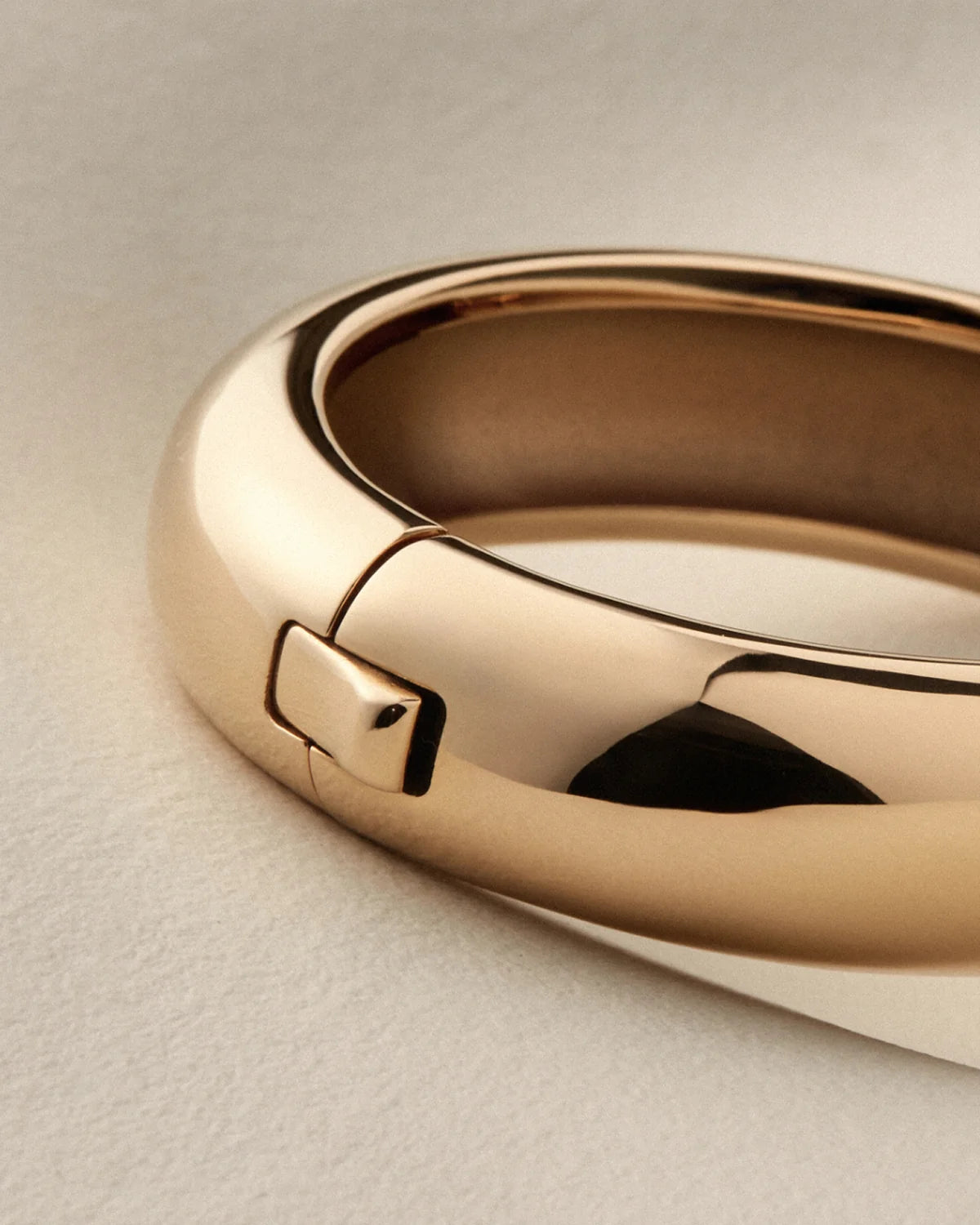 A close-up of a polished gold bangle bracelet with a sliding clasp, resting on a textured light beige surface.
