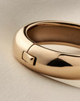 A close-up of a polished gold bangle bracelet with a sliding clasp, resting on a textured light beige surface.