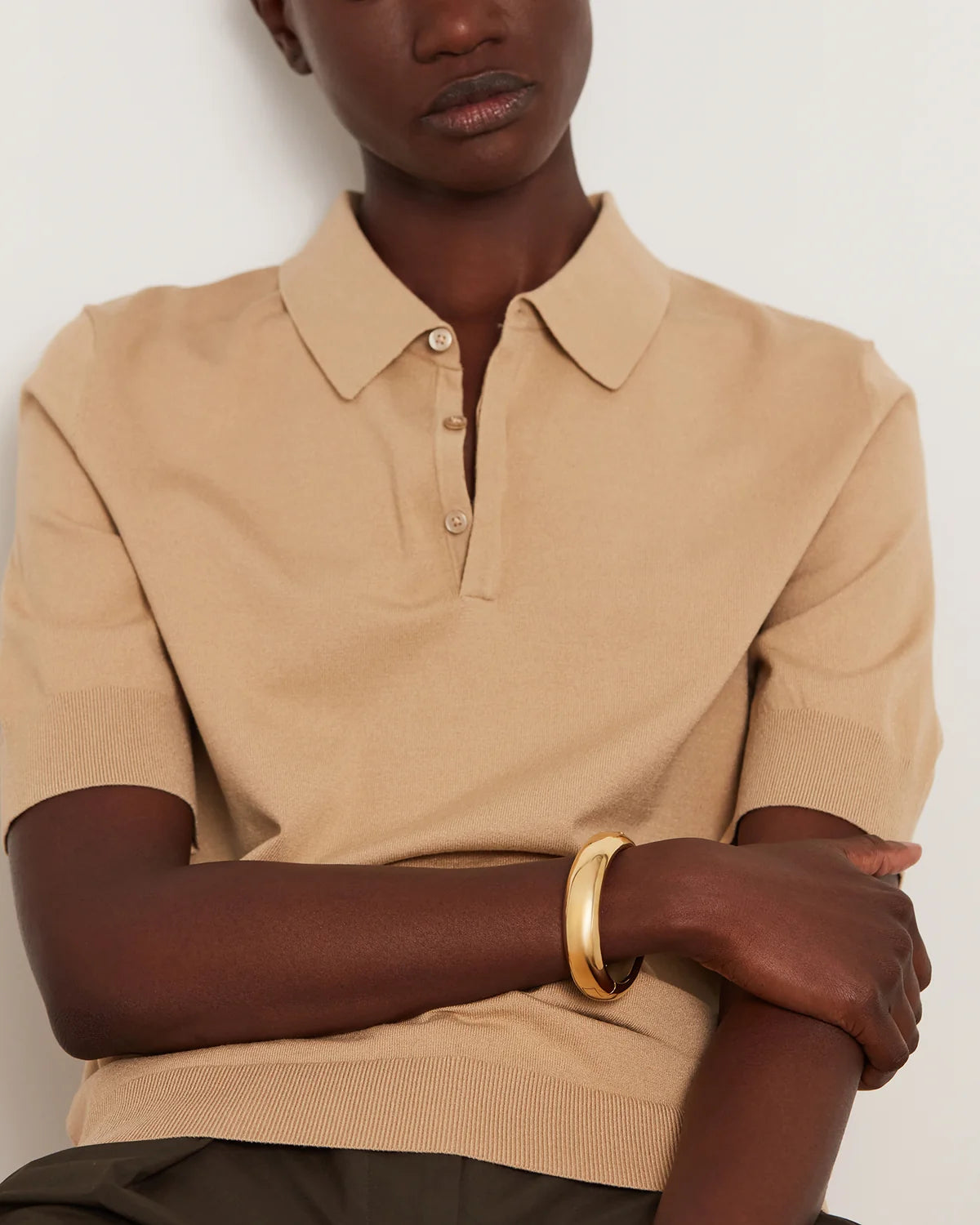A close-up of a model wearing a beige short-sleeve knit polo shirt with a buttoned collar, paired with a gold bangle bracelet, against a plain light background.