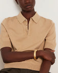 A close-up of a model wearing a beige short-sleeve knit polo shirt with a buttoned collar, paired with a gold bangle bracelet, against a plain light background.