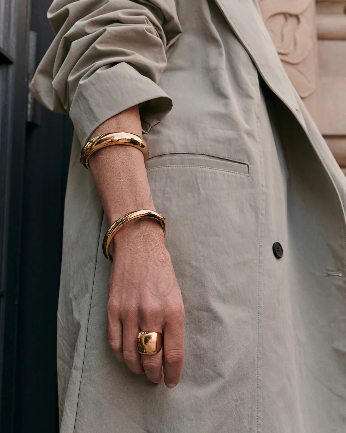 A close-up of a person wearing wide, polished gold bangle bracelets and a chunky gold ring, styled with a light beige trench coat.