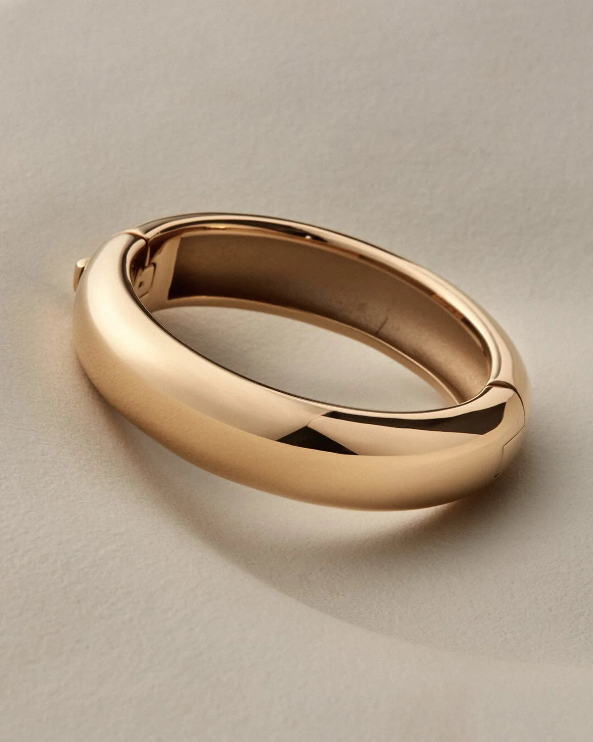 A polished gold bangle with a smooth, rounded design, resting on a neutral light beige surface.