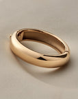 A polished gold bangle with a smooth, rounded design, resting on a neutral light beige surface.