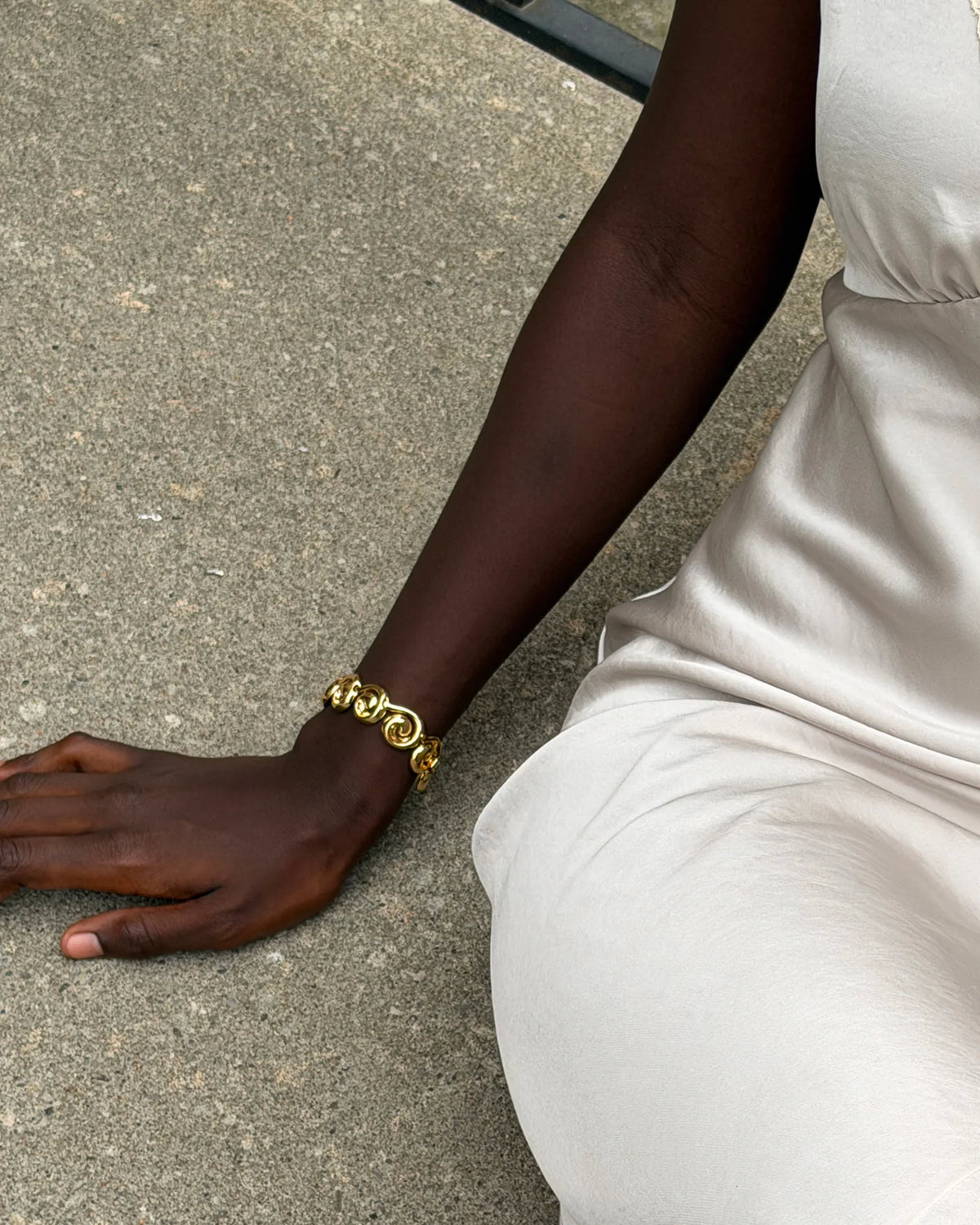 A close-up lifestyle shot of a person wearing a chunky gold chain link bracelet on their wrist, sitting on a concrete surface.