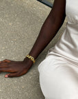 A close-up lifestyle shot of a person wearing a chunky gold chain link bracelet on their wrist, sitting on a concrete surface.