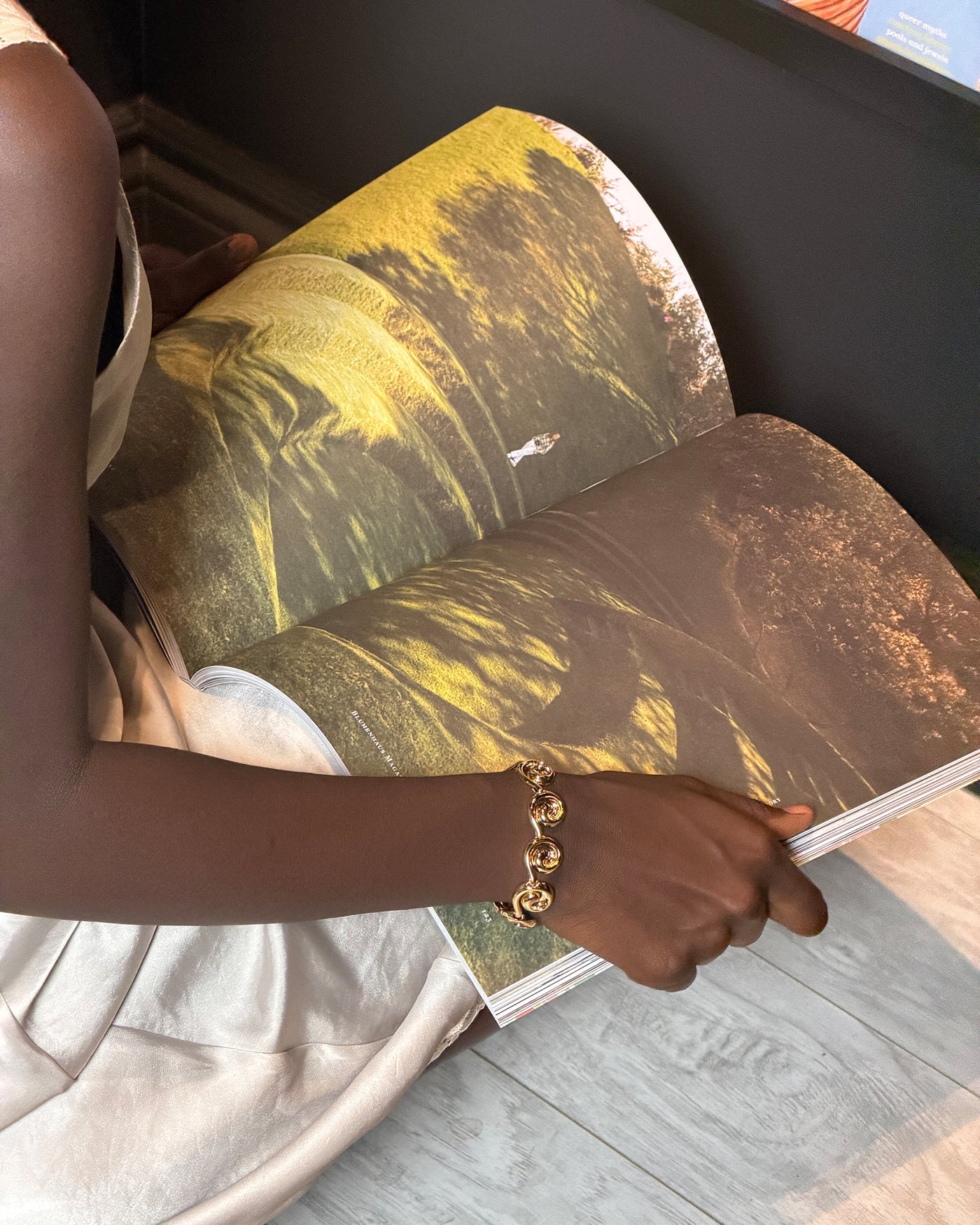 A person holding open a large printed book or magazine showing a landscape photograph with yellow and brown tones, wearing a gold chain bracelet on one arm.