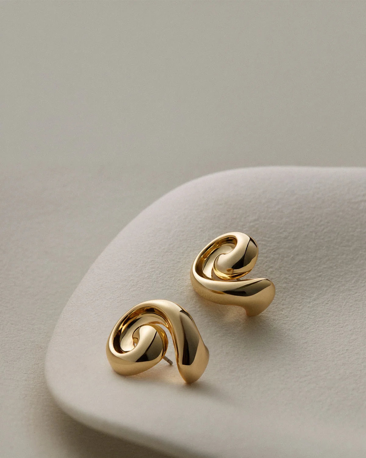 A pair of gold-toned abstract spiral stud earrings resting on a light beige textured display stand against a soft neutral background.