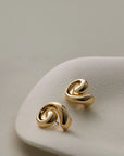 A pair of gold-toned abstract spiral stud earrings resting on a light beige textured display stand against a soft neutral background.