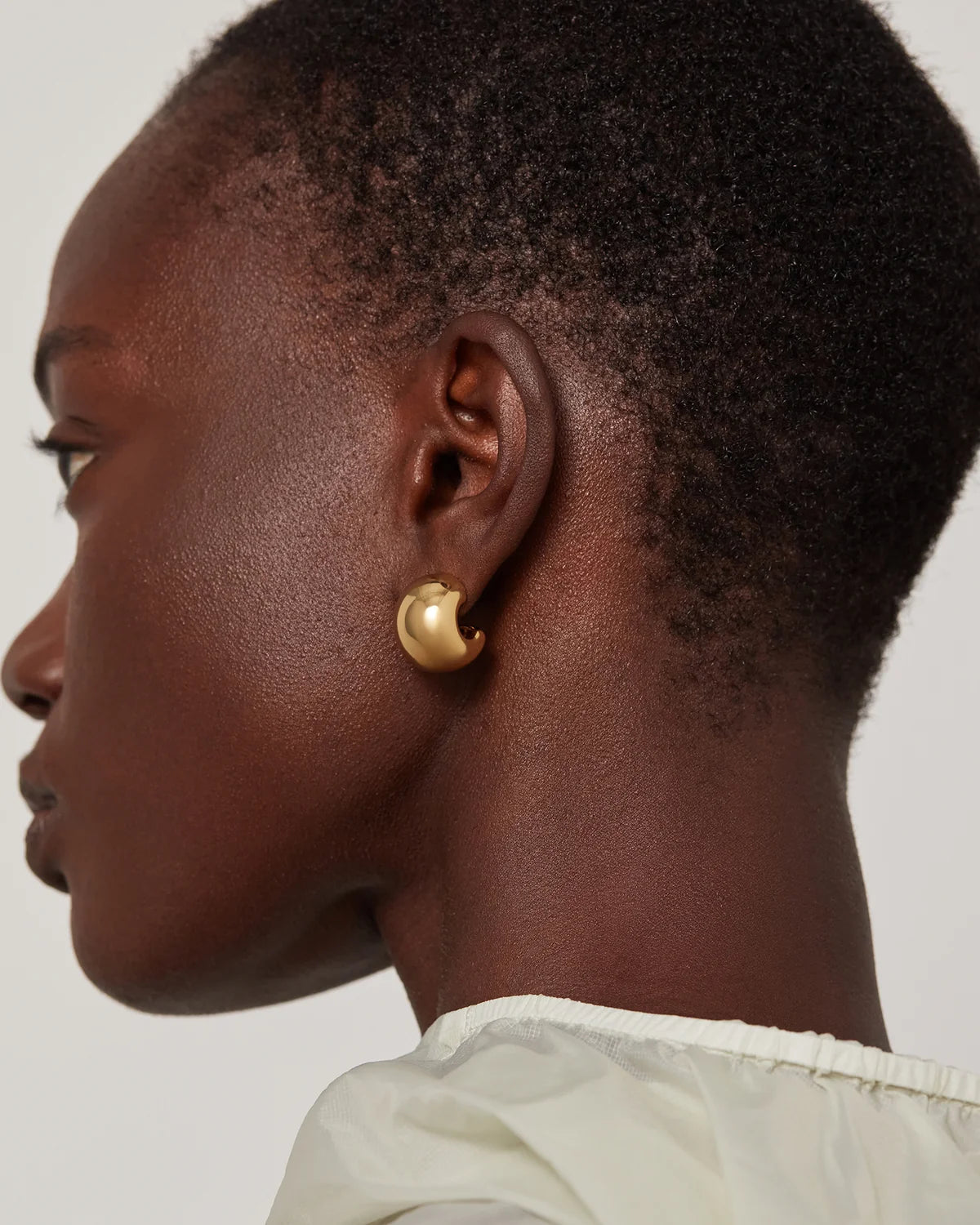 A close-up side profile of a person wearing a small, polished gold crescent-shaped stud earring against a light gray background.