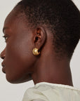 A close-up side profile of a person wearing a small, polished gold crescent-shaped stud earring against a light gray background.