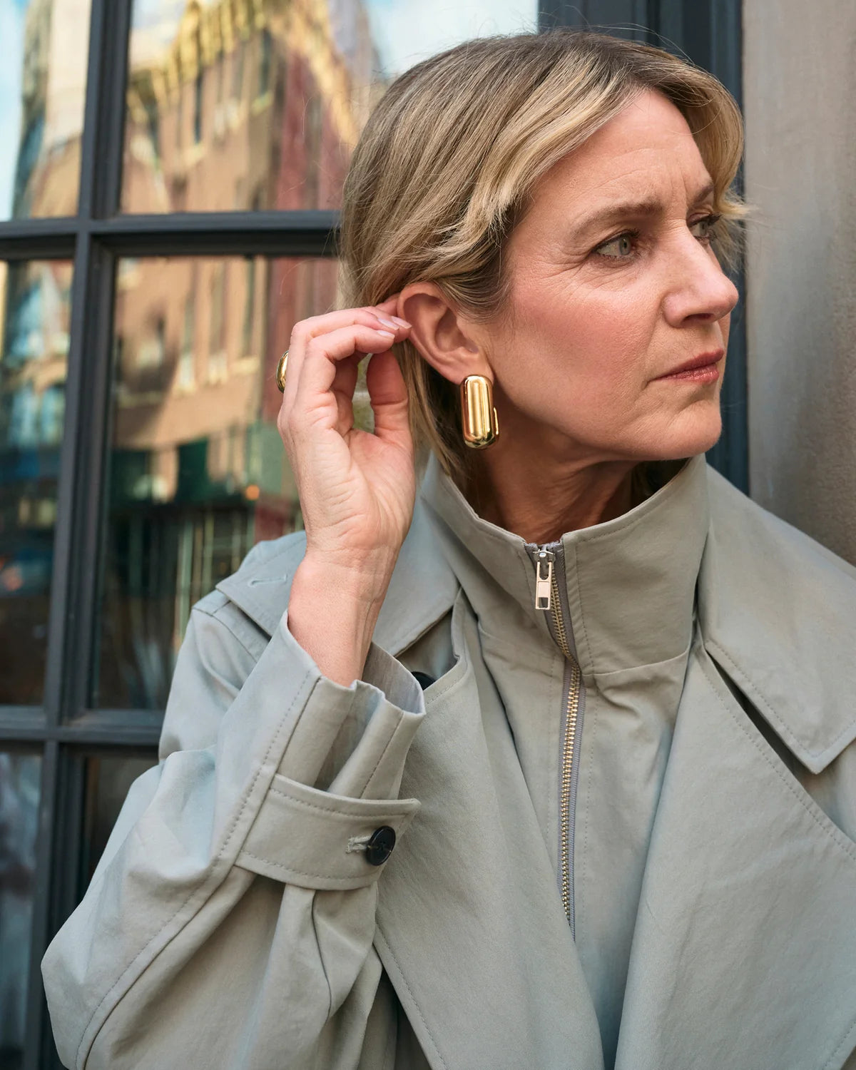 A woman wearing a light beige trench coat and gold geometric drop earrings, posing in front of a window with reflections.