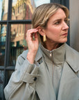 A woman wearing a light beige trench coat and gold geometric drop earrings, posing in front of a window with reflections.