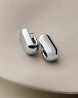 A pair of polished silver-toned geometric stud earrings with a smooth, rounded organic shape, displayed on a textured beige surface.