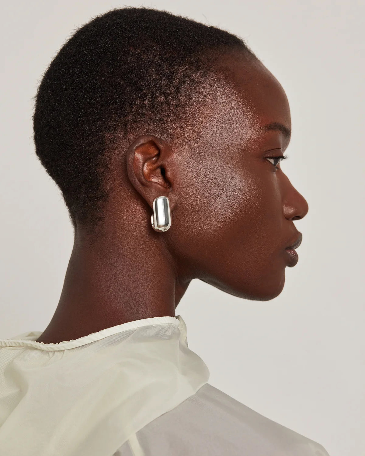 A close-up side profile of a model wearing a chunky, polished silver geometric drop earring against a light gray background.