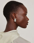 A close-up side profile of a model wearing a chunky, polished silver geometric drop earring against a light gray background.