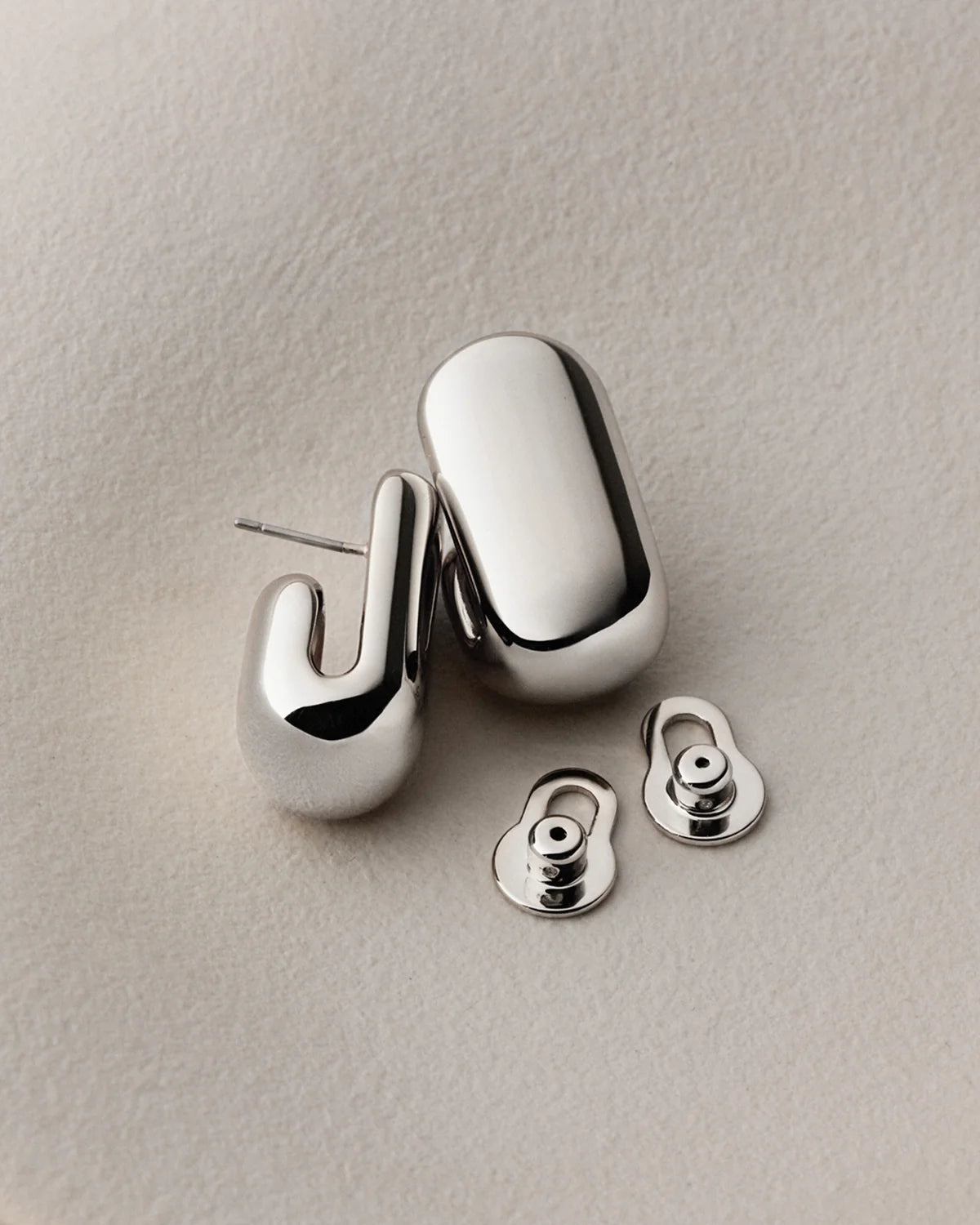 A pair of polished silver-toned geometric stud earrings with matching butterfly backings, displayed on a textured light beige surface.