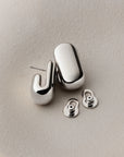 A pair of polished silver-toned geometric stud earrings with matching butterfly backings, displayed on a textured light beige surface.