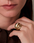 A close-up of a person wearing a large, sculptural gold ring on their finger, with their hand resting near their neck.