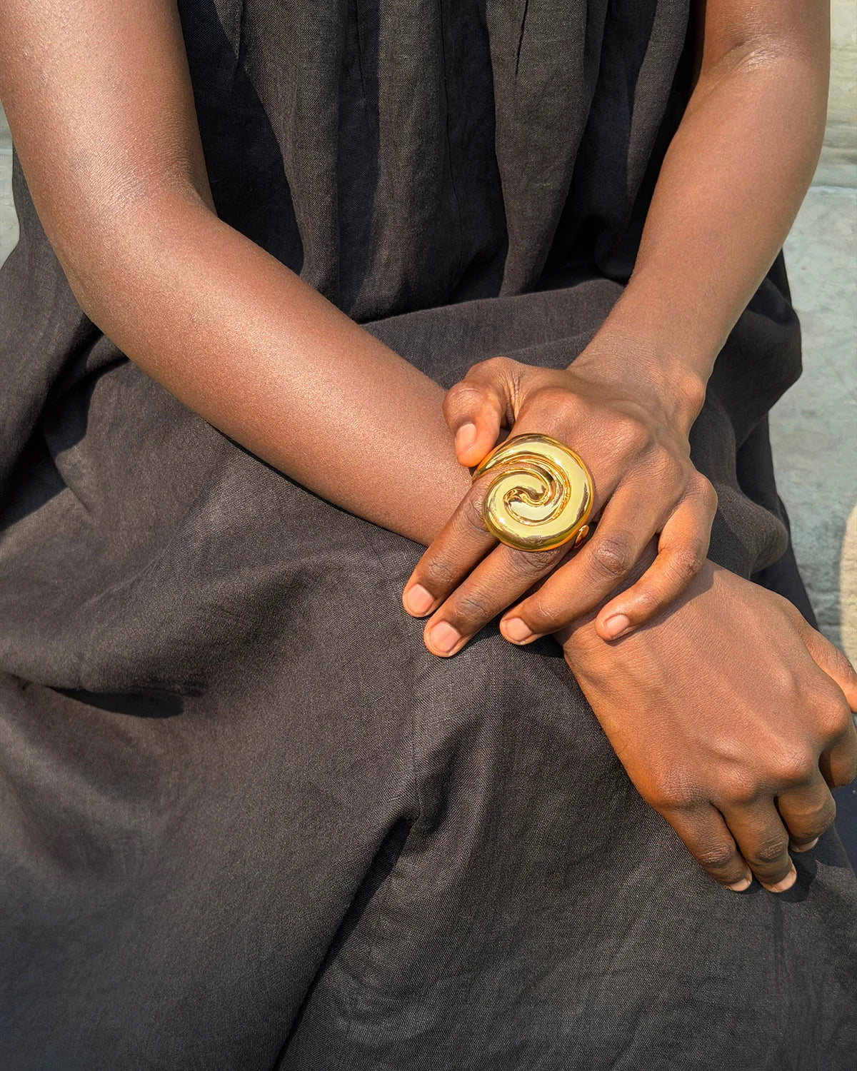 A close-up of a person wearing a large, chunky gold ring with a spiral design on their hand, dressed in a dark gray linen garment.