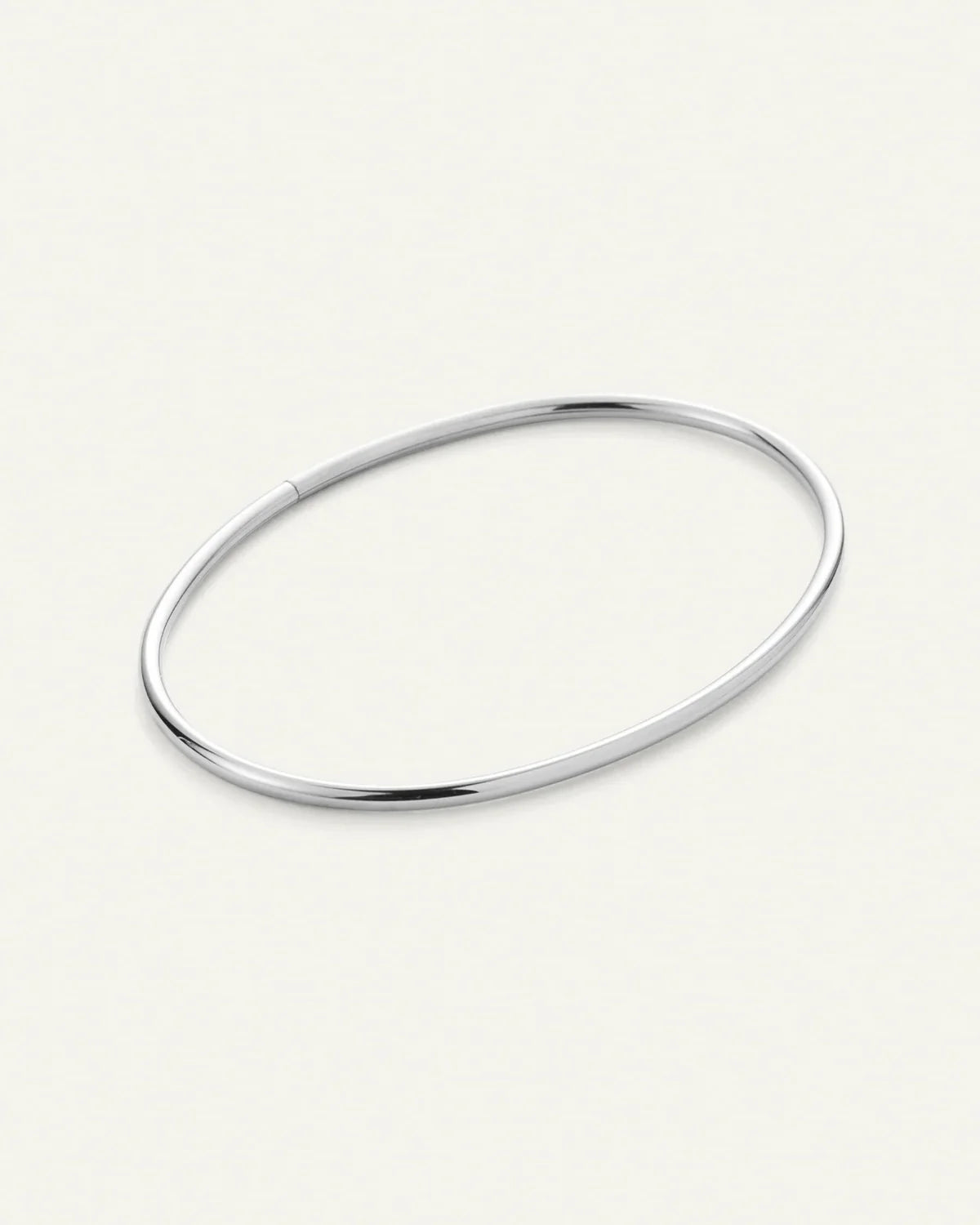 A minimalist, polished silver-toned metal bangle bracelet with a smooth, rounded oval shape, displayed on a clean, light cream background.