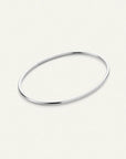 A minimalist, polished silver-toned metal bangle bracelet with a smooth, rounded oval shape, displayed on a clean, light cream background.
