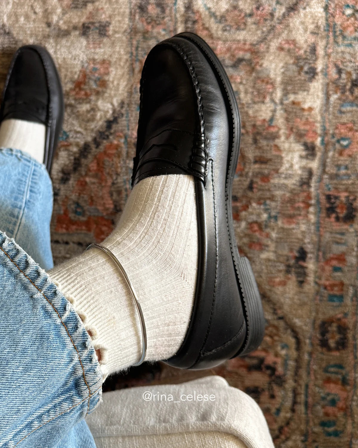 A person wearing black leather penny loafers with off-white ribbed socks and light blue jeans, standing on a patterned area rug. The brand name 'rina_celeste' is watermarked at the bottom.