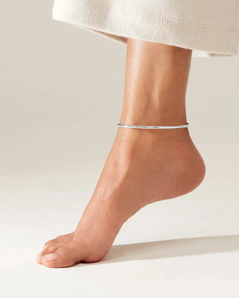A minimalist, thin silver anklet worn on a person's ankle, shown against a clean, light beige background with a portion of a white textured skirt visible at the top.