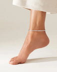 A minimalist, thin silver anklet worn on a person's ankle, shown against a clean, light beige background with a portion of a white textured skirt visible at the top.