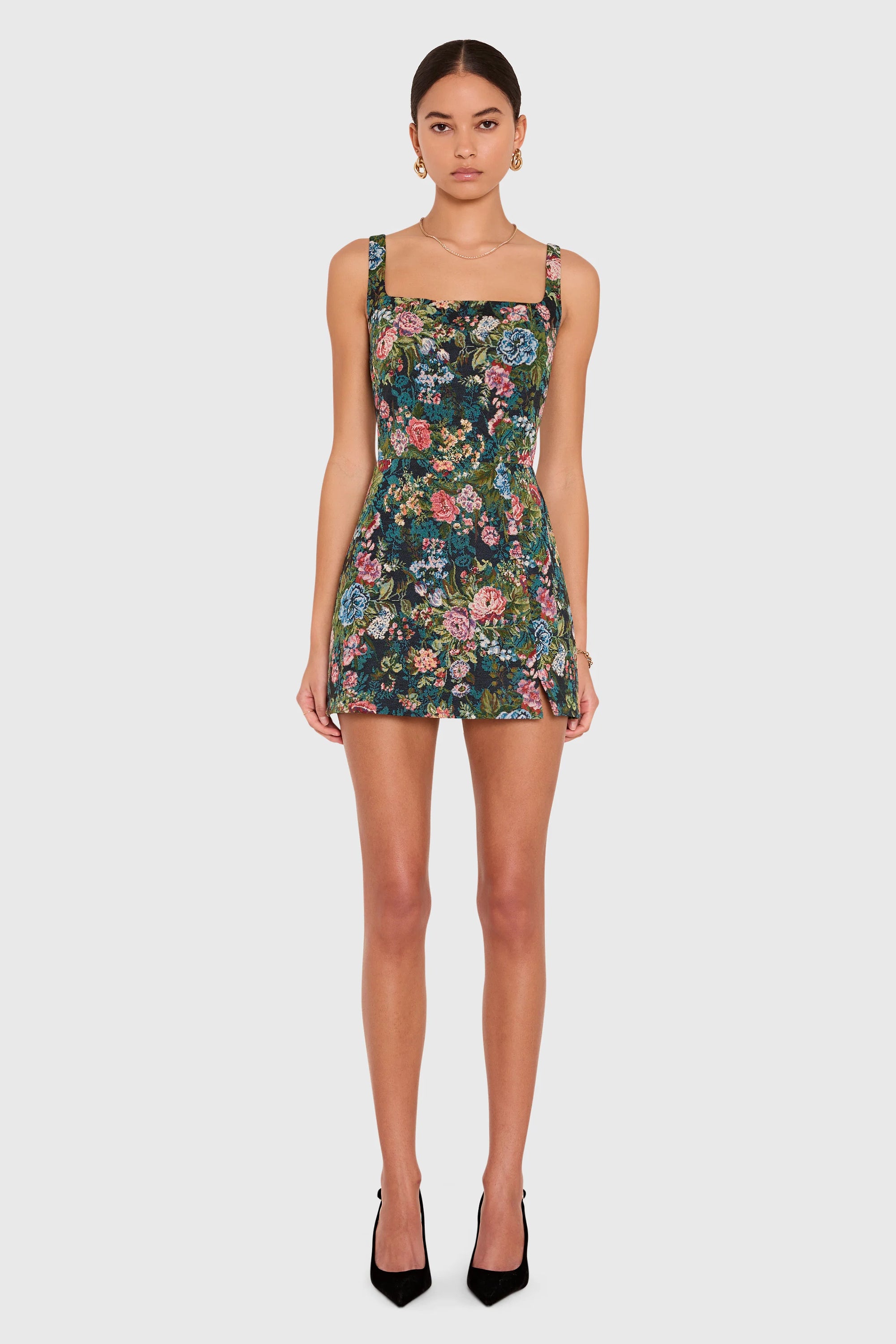 A model wearing a sleeveless mini dress with a dark green floral print, featuring a square neckline and thin shoulder straps, paired with black high heels against a light gray background.